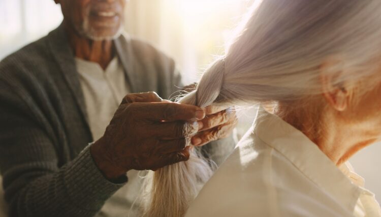An old man with shaky hands focuses on braiding long silver hair. He sits in a sunlit room with soft light filling the space. The scene captures a moment of care and attention.